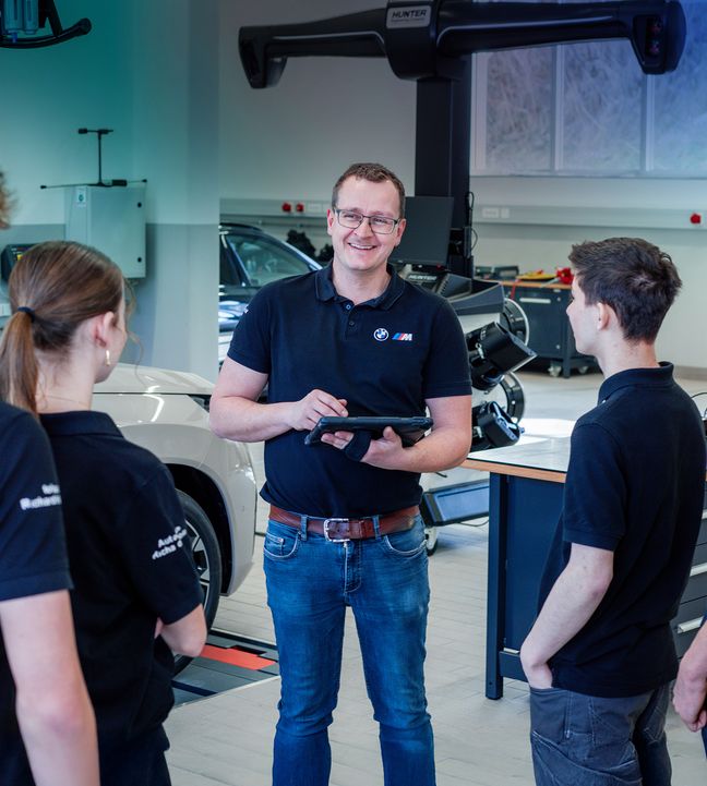 BMW-Techniker mit Tablet unterweist vier Auszubildende in moderner Kfz-Werkstatt, Diagnosegerät und Fahrzeuge im Hintergrund.